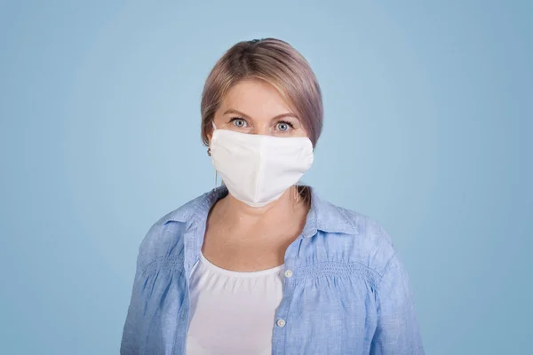 Mulher caucasiana sênior em t-shirt branca e camisa posando em uma parede azul usando uma máscara médica no rosto olhando para a câmera — Fotografia de Stock