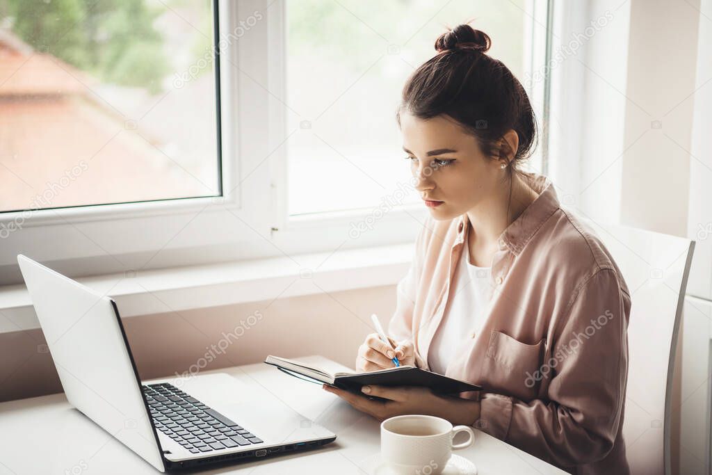 Concentrated caucasian woman in shirt writing a recipe in the book while looking at the laptop and drinking a cup of tea near the window