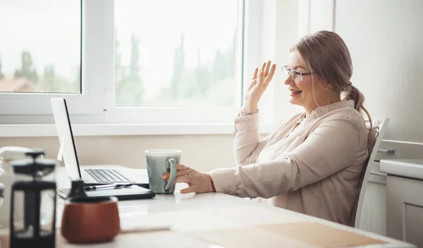 Senior mujer caucásica con anteojos bebiendo té y sonriendo a la computadora portátil gesto hola — Foto de Stock
