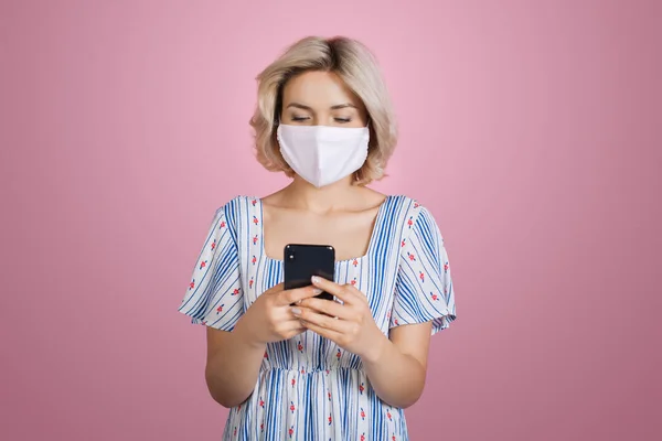 Wanita pirang yang cantik bertopeng medis mengenakan gaun bagus mengobrol di ponsel berpose di dinding studio merah muda — Stok Foto