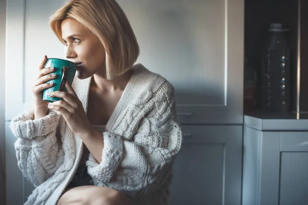 Krásná blondýnka s pleteným svetrem popíjející čaj u okna a usmívající se — Stock fotografie