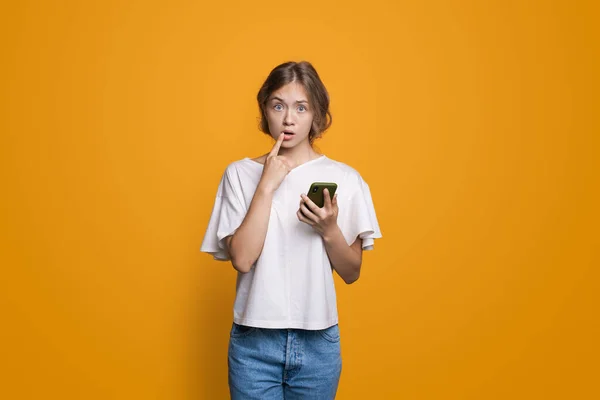 Mujer rubia caucásica pensativa sosteniendo un móvil y mirando a la cámara posando en una pared de estudio amarillo — Foto de Stock