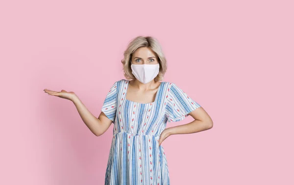 Wanita pirang Kaukasia yang mempesona mengiklankan sesuatu dengan topeng medis di dinding studio merah muda dengan gerakan ruang kosong dengan telapak tangan — Stok Foto