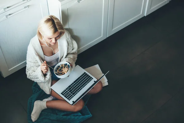 Foto vista superior de una mujer caucásica con cabello rubio y auriculares sentados en la cocina en el suelo y usando una computadora portátil comiendo cereales — Foto de Stock
