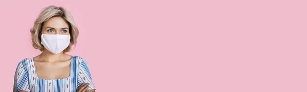 Piękna blondynka z maską medyczną na twarzy pozowanie na różowej ścianie studio z wolnej przestrzeni w ładnej sukience — Zdjęcie stockowe