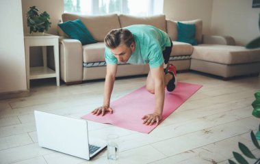 Diz üstü bilgisayarı olan sarışın, beyaz bir adam evde, yerde spor yapıyor.