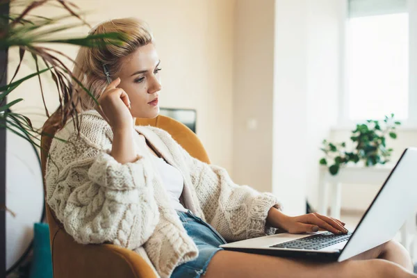 Mujer caucásica pensativa con el pelo rubio usando un ordenador portátil en casa usando un suéter de punto caliente — Foto de Stock