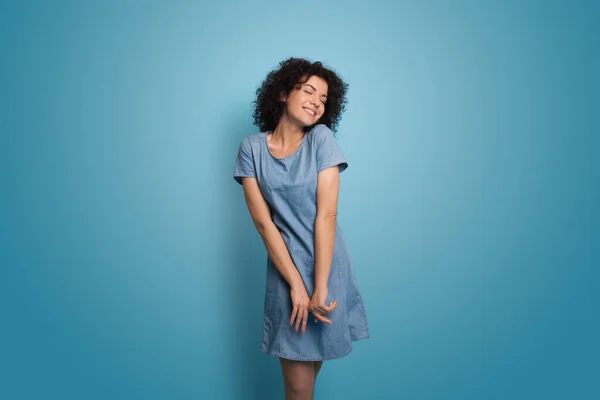 Deliciosa mujer caucásica con el pelo rizado usando un vestido azul de verano está sonriendo en una pared de estudio — Foto de Stock