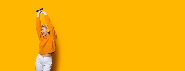 Blonde kaukasische Frau mit Kopfhörer hält ein Telefon in der Hand und hört Musik an einer gelben Studiowand mit freiem Platz in einem Pullover — Stockfoto