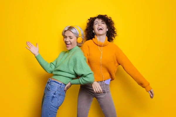 Zwei glückliche Schwestern mit lockigem Haar lachen, während sie Kopfhörer auf einer gelben Studiowand tragen — Stockfoto