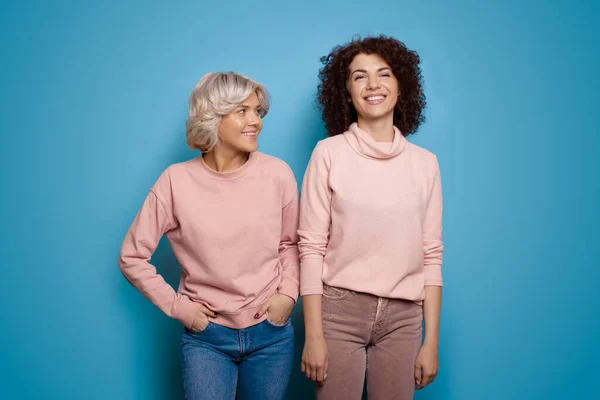 Charmante Frau mit lockigem Haar und ihre brünette Freundin lächeln in den gleichen Kleidern auf einer blauen Studiowand — Stockfoto