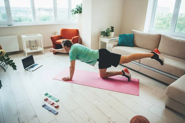 Вид збоку фото кавказького чоловіка в спортивному одязі, що тягнеться на підлогу, використовуючи ноутбук і килим вдома — стокове фото