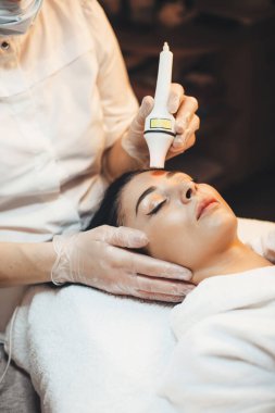 Spa salonunda yatan beyaz bir kadının modern aletlerle yüz yaşlanmasına karşı prosedür uyguladığını gösteren yan görüntü fotoğrafı.