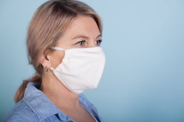 Yüzüne tıbbi maske takan beyaz bir kadın mavi stüdyo duvarında poz veriyor.