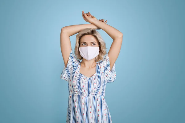 Blonde Dame mit medizinischer Maske im Gesicht und Kleid posiert mit den Händen oben auf blauer Studiowand — Stockfoto