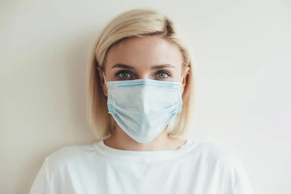 Photo de face d'une femme caucasienne aux cheveux blonds regardant la caméra et portant un masque médical sur le visage sur un fond de studio blanc — Photo
