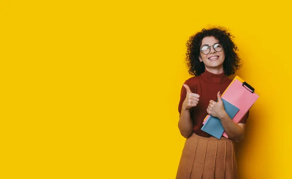 Charming brunette lady with curly hair and eyeglasses holding some folders and gesturing the like sign on a yellow studio wall with free space — Stock Photo, Image