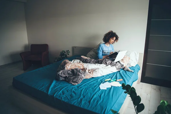 Wanita berambut keriting membaca buku di pagi hari duduk di tempat tidur dengan laptop — Stok Foto