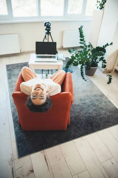 Jovem blogueiro de vídeo sentado em poltrona e sorrir usando óculos na frente de um laptop com câmera — Fotografia de Stock