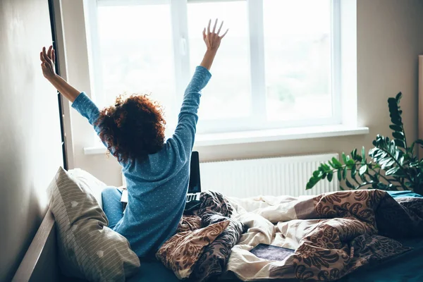 Mujer caucásica de pelo rizado en pijama azul estirándose en la cama temprano en la mañana —  Fotos de Stock