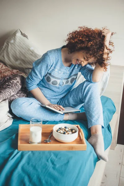 Sonriente chica caucásica con el pelo rizado leyendo un libro en la cama y comiendo cereales saludables con leche — Foto de Stock