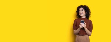 Kıvırcık saçlı esmer kadın telefonda konuşuyor ve sarı stüdyo duvarına gülümsüyor.