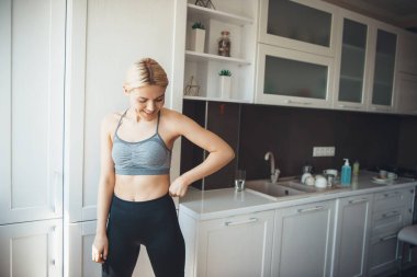 Spor giyimli beyaz kadın mutfakta gülümsüyor.