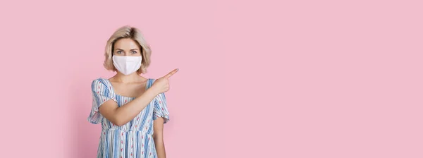 Blanke vrouw met blond haar draagt een medisch masker op gezicht wijst naar roze vrije ruimte in de buurt van haar — Stockfoto