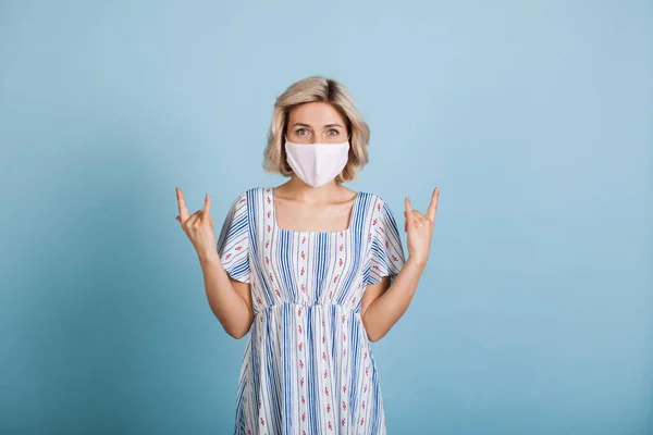 Schöne blonde Frau mit medizinischer Maske auf dem Gesicht gestikuliert das Felsschild auf einer blauen Studiowand — Stockfoto