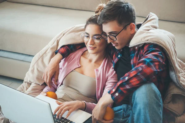 Fiatal kaukázusi pár ül a padlón egy takaróval borított kezében egy narancs és nézi a laptop — Stock Fotó