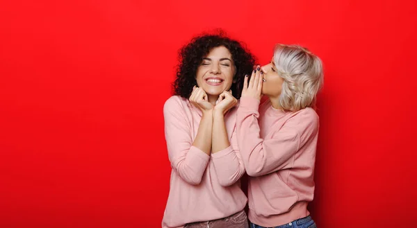 Lockige blonde Frau flüstert ihrem brünetten Freund etwas zu und träumt von einer roten Studiowand — Stockfoto