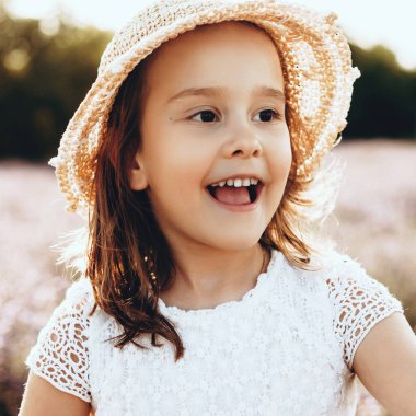 Tarlada poz verirken gülümseyen şapkalı ve elbiseli neşeli küçük kız.