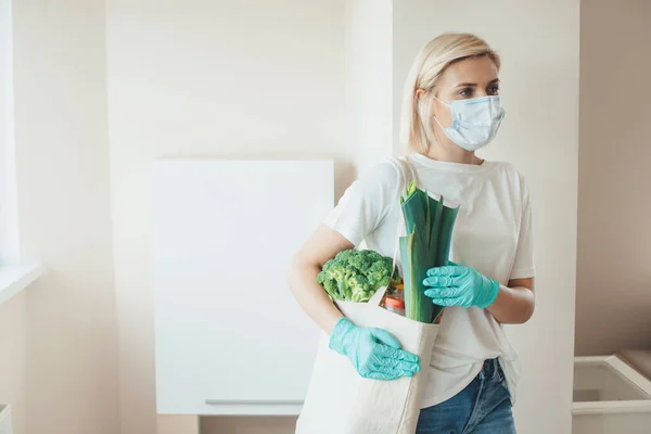 Blonde kaukasische Frau mit medizinischer Maske im Gesicht und Handschuhen hält eine Tasche mit Produkten — Stockfoto