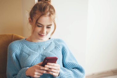 Çilli kızıl kadın, evde mavi örülü bir süveter giyip cep telefonuyla sohbet ediyor.
