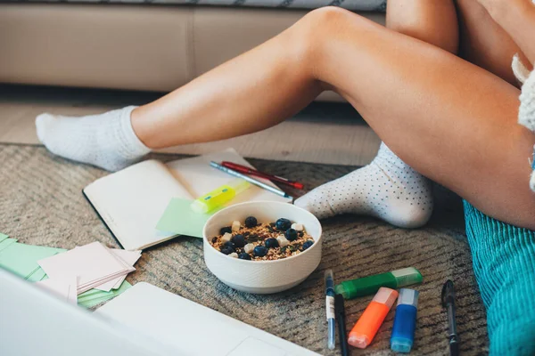 Mujer caucásica sentada en el suelo y comiendo cereales con bayas mientras hace los deberes durante el encierro — Foto de Stock