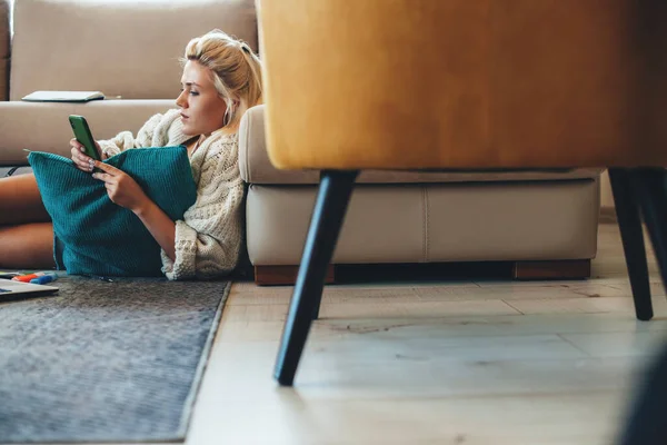 Mujer rubia con un suéter de punto usando un teléfono en el suelo mientras trabaja en el portátil — Foto de Stock