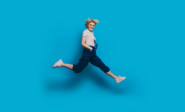 Enérgica mulher encantadora com cabelo loiro sorrindo para a câmera e pulando em uma parede de estúdio azul — Fotografia de Stock