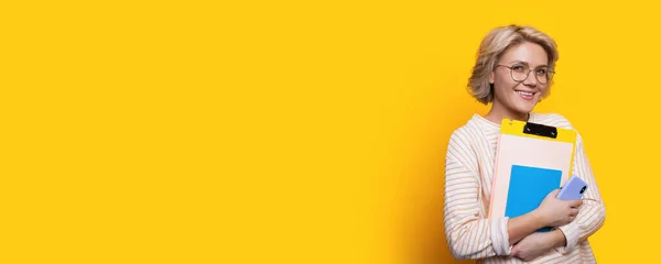 Charming blonde student with eyeglasses holding some folders smiling at camera on a yellow wall with free space — ストック写真