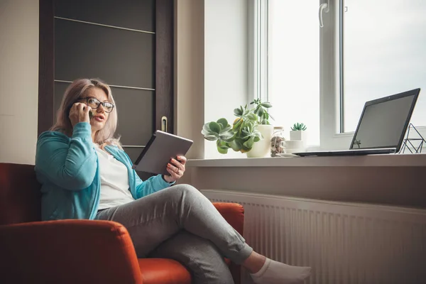 Busy senior woman with blonde hair and eyeglasses holding a tablet and talking on phone while working from home — Stock Photo, Image