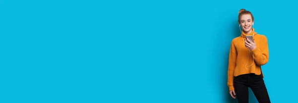 Mujer de jengibre caucásico con pecas en un suéter amarillo está escuchando a meditar a través de auriculares y móviles en una pared azul con espacio libre — Foto de Stock