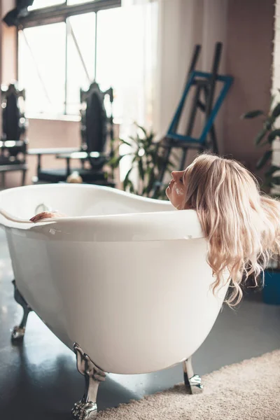 Ανώτερος καυκάσιος γυναίκα με ξανθά μαλλιά χαλαρώνοντας σε μια μπανιέρα στο σπίτι, ενώ βάζοντας στο πρόσωπο μια αντιγηραντική κρέμα — Φωτογραφία Αρχείου