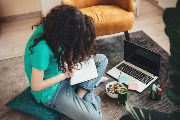Översiktsbild av en kaukasisk student som gör läxor på golvet och använder en bärbar dator medan du äter spannmål med grönsaksjuice — Stockfoto