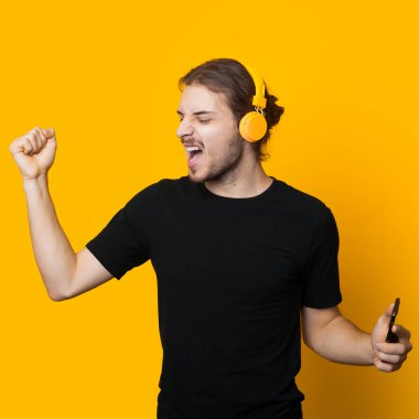 Siyah tişörtlü beyaz bir adam, kulaklıkla müzik dinliyor ve sarı bir stüdyo duvarında dans ediyor.