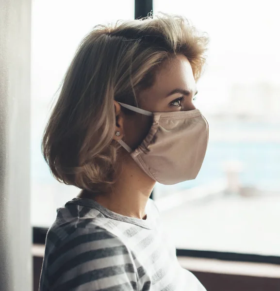 Foto vista lateral de una mujer caucásica con cabello rubio y máscara médica en la cara mirando en algún lugar cerca de la ventana — Foto de Stock