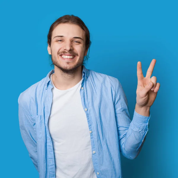 Toothily sorrindo homem com barba e cabelos longos gesticulando paz e amizade em uma parede de estúdio azul — Fotografia de Stock