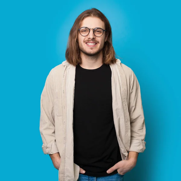 Homem caucasiano com cabelos longos e barba vestindo óculos está sorrindo para a câmera em uma parede de estúdio azul — Fotografia de Stock