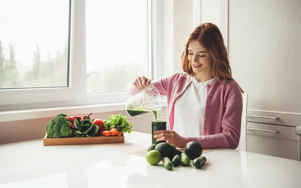 Wanita berambut merah dengan bintik-bintik menaruh beberapa jus sayuran hijau di kaca dan tersenyum dekat beberapa buah-buahan — Stok Foto