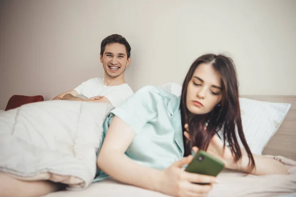 Sonriente hombre caucásico esperando a su esposa para escribir un mensaje en el teléfono en la cama —  Fotos de Stock