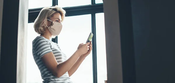 Foto vista lateral de una mujer caucásica con el pelo corto y rubio usando una máscara médica en la cara está usando un teléfono cerca de la ventana — Foto de Stock