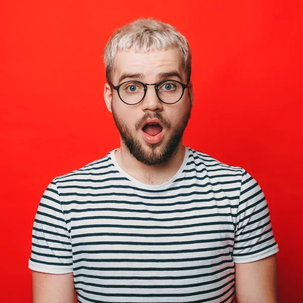 Blondynka biały mężczyzna gestykulując zdumienie podczas noszenia okularów na czerwonej ścianie studio — Zdjęcie stockowe